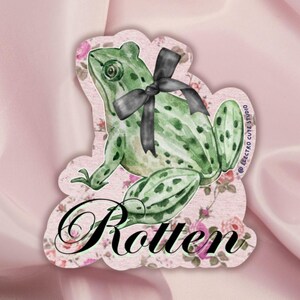 Puede incluir: Una rana verde con un lazo negro sobre un fondo rosa con patrones florales. La palabra "Rotten" está escrita en letras cursivas negras.