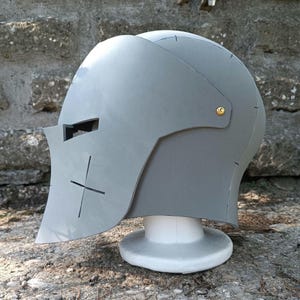 Könnte beinhalten: Ein grauer Helm mit einem Kreuzmuster auf der Vorderseite und einem goldenen Knopf an der Seite. Der Helm ist aus Schaumstoff und steht auf einem weißen Ständer.
