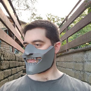 Könnte beinhalten: Eine Person trägt eine graue, detaillierte Maske, die Mund und Nase bedeckt. Die Maske hat ein scharfes, kantiges Design mit einem markanten, zahnigen Grinsen.