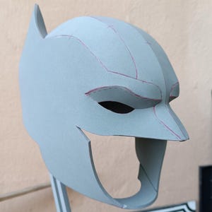 Könnte beinhalten: Eine hellgraue Maske im Batman-Stil, hergestellt aus einem starren Material. Die Maske hat spitze Ohren, Augenöffnungen und eine definierte Nase. Rosa Linien markieren die Konturen des Designs. Die Maske ist auf einem Metallständer montiert.