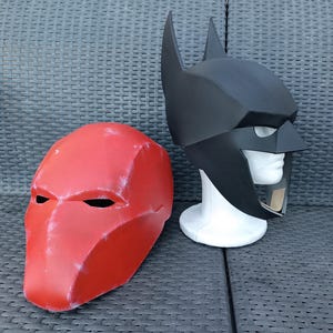 May include: Two superhero masks, one red and one black. The red mask has a more angular design, while the black mask has a more rounded design. Both masks are made of a hard plastic material.