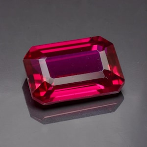 Puede incluir: Una gema facetada de corte rectangular con un tono magenta intenso. Las facetas de la gema reflejan la luz, creando una brillante exhibición de color. La gema se encuentra sobre un fondo oscuro, resaltando su color vibrante y claridad.