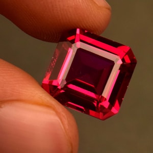 May include: A deep red, square-shaped gemstone with a faceted cut.