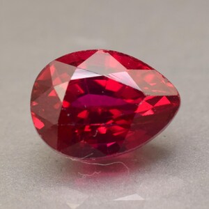 May include: A pear-shaped, faceted gemstone with a deep, vibrant red color. The stone features a symmetrical cut with multiple triangular and trapezoidal facets that reflect light, creating a brilliant, sparkling appearance across its surface.