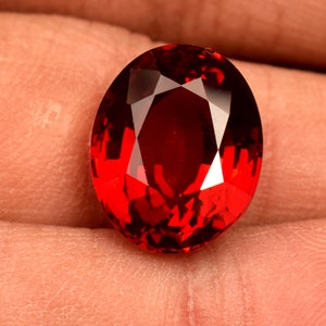 May include: A deep red oval-shaped gemstone with a faceted cut. The gemstone is held in a hand.