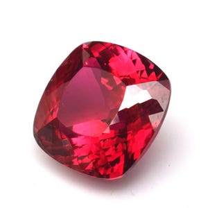 Könnte beinhalten: Ein facettierter Edelstein im Kissenschliff mit tiefroter Farbe. Der Stein hat eine hohe Klarheit und viele Facetten, die das Licht reflektieren. Die Form ist ein modifiziertes Quadrat mit abgerundeten Ecken, was einen lebendigen Eindruck erzeugt.
