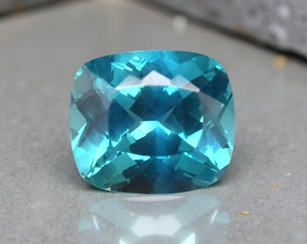 May include: A faceted, cushion-cut gemstone in shades of turquoise and teal. The gem's surface reflects light, showcasing its intricate cuts and internal structure. The gemstone is set against a neutral background, highlighting its vibrant color and clarity.