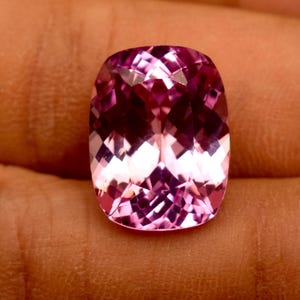 Könnte beinhalten: Ein facettierter, kissenförmiger Edelstein in Rosa. Der Edelstein hat eine komplexe innere Struktur, die das Licht reflektiert und einen funkelnden Effekt erzeugt. Der Stein hat eine rechteckige Form mit abgerundeten Ecken und wird in einer Hand gehalten.