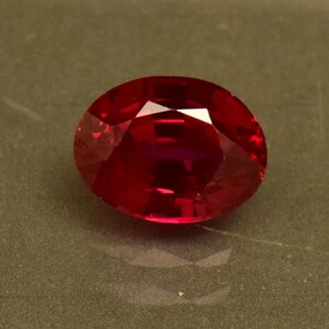 May include: A vibrant red oval-shaped gemstone with a faceted cut. The gemstone is set against a neutral background.