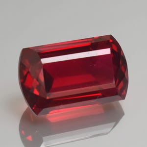 May include: A rectangular-cut, faceted gemstone with a deep red hue. The gem has a polished surface with sharp edges and a slight curve on the ends. The gem is displayed on a reflective surface, showcasing its brilliance.