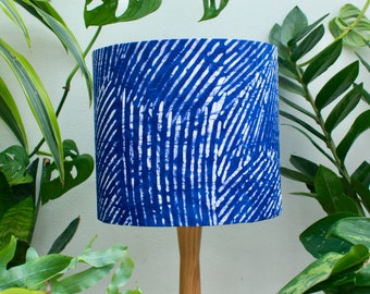 May include: A cylindrical lampshade with a vibrant blue and white pattern. The design features diagonal white lines against a deep blue background. The lampshade is mounted on a wooden base, surrounded by lush green plants.
