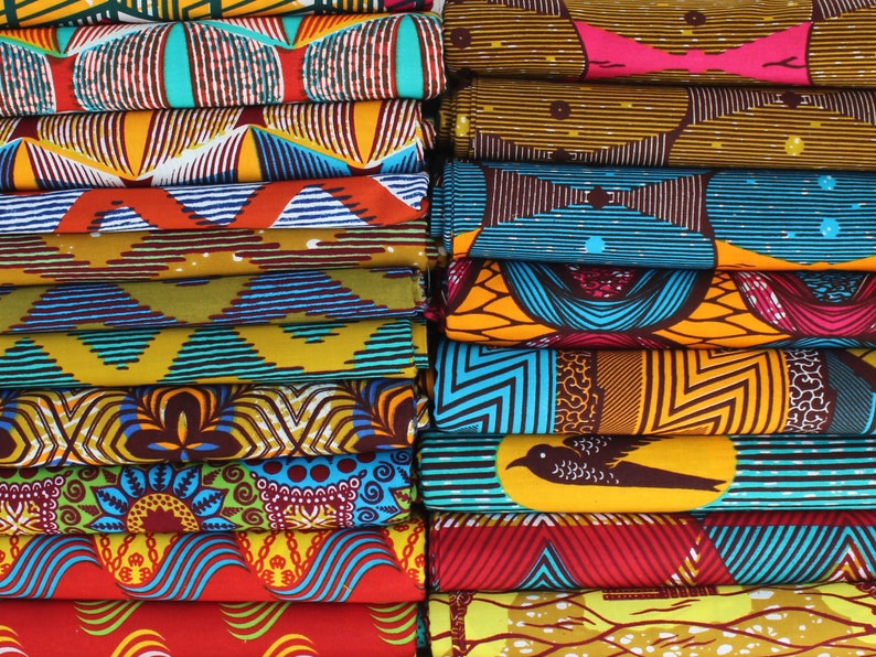 May include: A stack of colorful fabric with various patterns, including geometric designs, floral motifs, and abstract shapes. The fabric is folded neatly and arranged in a vertical stack.