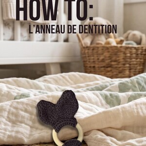 Peut inclure: Un anneau de dentition composé d'un cercle en bois clair lisse orné de tissu crocheté gris anthracite en forme d'oreilles. Le texte "HOW TO: L'ANNEAU DE DENTITION" est affiché en lettres noires grasses en haut de l'image.