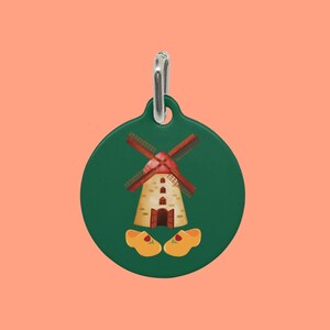 May include: A green circular tag with a silver loop. The tag features a cartoon illustration of a Dutch windmill with red blades and a yellow and brown building. Below the windmill are two yellow wooden shoes.