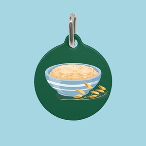 May include: A green, round pet tag featuring a bowl of oatmeal with blue stripes and a sprig of wheat. The oatmeal is a light tan color, and the tag has a silver-colored metal loop for attaching to a collar.