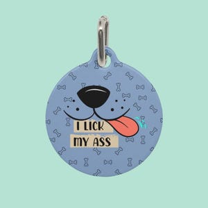 May include: A round, light blue pet tag with a dog face design and the text "I LICK MY ASS". The tag features a black nose, a red tongue, and a pattern of white dog bones. A silver metal ring is at the top.