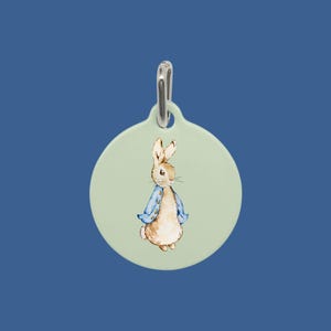 May include: A round, light green pet tag with a watercolor illustration of Peter Rabbit wearing a blue jacket. The tag has a silver-colored metal loop at the top for attaching to a collar. The background is a solid, dark blue.