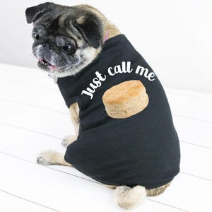 May include: A pug wearing a black dog shirt with the text "Just call me" and an image of a biscuit. The dog is sitting on a white surface and looking to the left.
