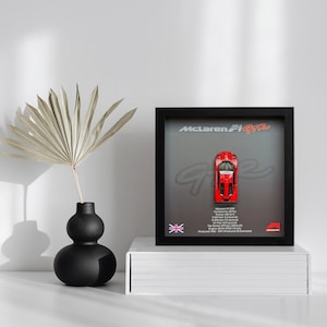 Puede incluir: Una vitrina negra enmarcada con un modelo de coche rojo McLaren F1 GTR en su interior. La vitrina tiene un fondo gris con el texto "McLaren F1 GTR" y una pequeña bandera del Reino Unido.