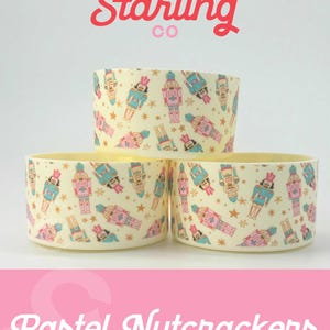 May include: Three pastel-colored ceramic containers with a nutcracker pattern. The containers are stacked on top of each other. The containers are decorated with gold stars and a white background. The containers are labeled "Sweet Starling Co. Pastel Nutcrackers".