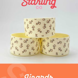 May include: Three yellow ceramic bowls with a pattern of gray lizards. The bowls are stacked on top of each other. The top bowl has the text "Sweet Starling Co." in pink and red lettering. The bottom bowl has the text "Lizards" in red lettering.