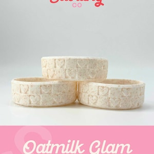 May include: Three off-white, circular containers with a heart-shaped brick pattern. The containers are stacked, with the text "Sweet Starling Co" in red above them. Below, the text "Oatmilk Glam PIXIE" is displayed on a pink background.