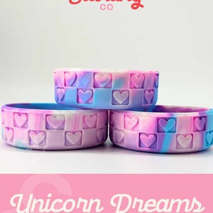 May include: Three stacked, round, silicone bracelets with a heart pattern. The bracelets are in shades of blue, pink, and purple, with the text "Sweet Starling Co" and "Unicorn Dreams Pixie" in the image.