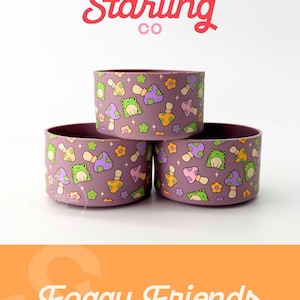 May include: Three purple silicone cups with a pattern of green frogs, mushrooms, and flowers. The cups are stacked on top of each other. The text "Foggy Friends" is written in white on an orange background.