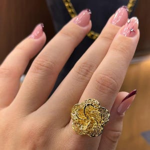 Op de afbeelding: Een goudkleurige ring met een bloemmotief. De ring heeft een opengewerkt patroon, waardoor hij een delicate, getextureerde uitstraling heeft. De ring wordt aan een vinger gedragen en benadrukt de ingewikkelde details en het vakmanschap.