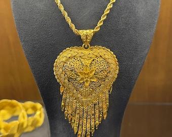 Może przedstawiać: Złoty naszyjnik z dużym, ozdobnym wisiorkiem w kształcie serca. Wisiorek ma skomplikowane filigranowe zdobienia i kwiatowy wzór pośrodku, z wiszącymi frędzlami. Naszyjnik jest prezentowany na szarej figurze.