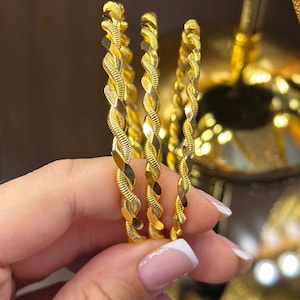 Puede incluir: Tres brazaletes de oro con un diseño de cuerda trenzada y acabado pulido. Cada brazalete está construido con un hilo metálico continuo que crea un patrón en espiral, reflejando la luz sobre la superficie texturizada de las piezas de joyería.