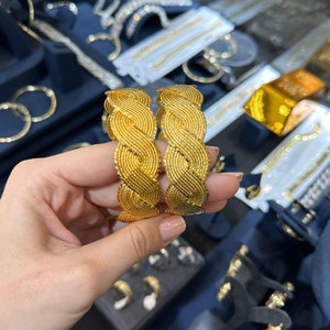 May include: Two matching gold bangles featuring a textured, braided, or woven pattern. The bracelets have a bright yellow gold finish with a fine, intricate surface detail that creates a repeating wave-like design across the entire circumference.
