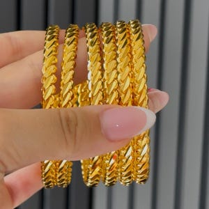 May include: A set of five gold bangles, each featuring a continuous, repeating braided or twisted link pattern. The bracelets have a highly reflective, polished finish that catches the light, creating a bright, metallic yellow appearance.