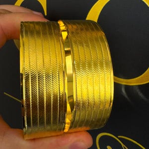 May include: Two wide, polished gold-tone cuff bracelets featuring a textured, repeating herringbone pattern across the surface. The bracelets have a smooth, reflective central band and smooth edges, showcasing a bright, metallic yellow finish.