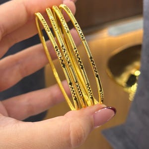 May include: A set of four thin gold bangles, each featuring a repeating geometric pattern of small etched triangles and fine horizontal lines along the outer surface. The bangles have a polished yellow gold finish and a slender, circular profile.