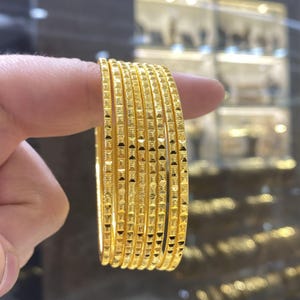 May include: A set of eight gold bangles, each with a repeating square pattern. The bangles are held between fingers, showcasing their bright, reflective surface. The background is blurred, suggesting a jewelry store setting.