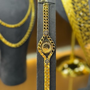 May include: A gold bracelet featuring a coin-shaped central element and detailed chain links. The bracelet is presented on a dark grey stand, highlighting its shiny surface. Other gold jewellery items are visible in the background.
