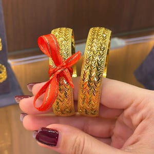Könnte beinhalten: Zwei goldene Armreifen mit einem sich wiederholenden, chevron- oder blattartigen Strukturmuster auf der Oberfläche. Einer der Armreifen ist mit einer kleinen, leuchtend roten Schleife verziert, die einen farblichen Kontrast zum Gold bildet.
