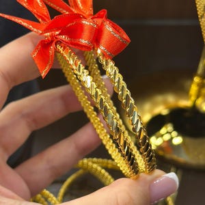 Può includere: Braccialetti color oro con un design strutturato sono tenuti in mano. Un nastro rosso con finiture dorate è legato attorno ai braccialetti. I gioielli sono esposti su uno sfondo scuro.