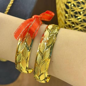 Könnte beinhalten: Zwei goldene Armreifen mit einem geprägten Blattmuster und einer strukturierten Oberfläche. Ein Armreif ist mit einer leuchtend roten Schleife verziert. Die Armreifen haben eine polierte, reflektierende Oberfläche und ein breites, gebogenes Design.