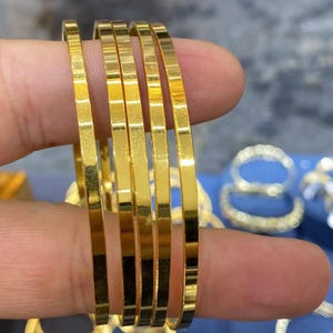 Peut inclure: Un ensemble de quatre joncs fins en métal doré poli avec un profil plat à bords carrés. Les bracelets présentent une surface métallique lisse et hautement réfléchissante qui capte la lumière, créant un aspect brillant et uniforme sur chaque anneau.