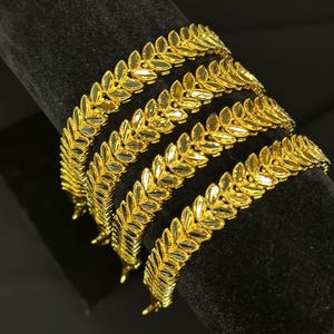 May include: Four gold leaf-shaped bangles stacked on a black velvet surface. The bangles are made of a shiny gold metal and have a delicate, intricate design.