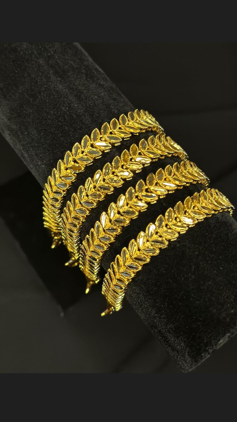 May include: Four gold leaf-shaped bangles stacked on a black velvet surface. The bangles are made of a shiny gold metal and have a delicate, intricate design.