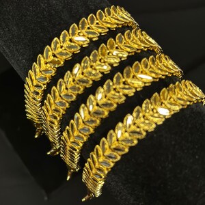 May include: Four gold bangles with a leaf design. The bangles are stacked on top of each other and are on a black background.