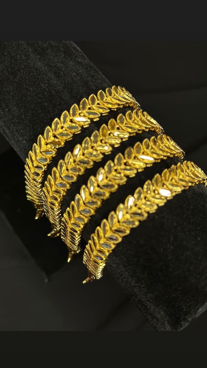 May include: Four gold bangles with a leaf design. The bangles are stacked on top of each other and are on a black background.
