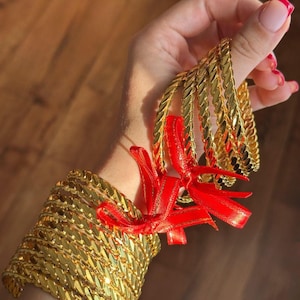 May include: A set of gold-colored metal bangles featuring a braided or twisted chain-link texture. The bangles are bundled together with a bright red ribbon tied in a bow, creating a decorative presentation for the collection of metallic bracelets.