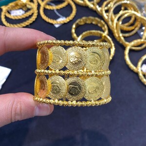 May include: A wide gold cuff bracelet featuring two horizontal rows of circular coin-like medallions. The edges of the bracelet are lined with textured, beaded gold wire, creating a decorative border around the central coin design.