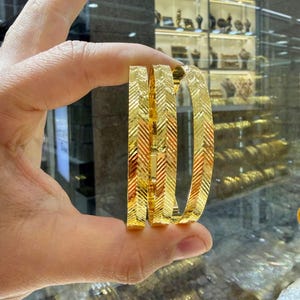 Pode incluir: Três pulseiras de ouro com um padrão de espinha de peixe diagonal repetitivo gravado na superfície metálica. As pulseiras têm um acabamento polido e um perfil plano e estreito, refletindo a luz nas linhas texturizadas e anguladas do exterior.