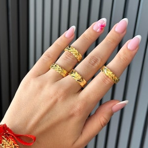 Op de afbeelding: Vier gouden ringen met een herhalend blad- of visgraatpatroon gedragen aan de vingers, vergezeld van een bijpassende gouden armband met een rood koordje. De ringen en armband hebben een gepolijste geelgouden afwerking met textuur.