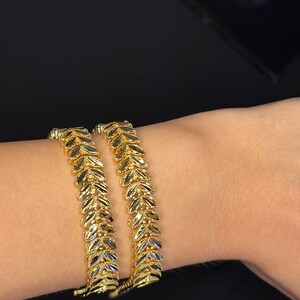 May include: Two gold leaf-shaped bangles on a wrist. The bangles are made of two-toned gold, with a lighter gold color on the inside of the leaves and a darker gold color on the outside.